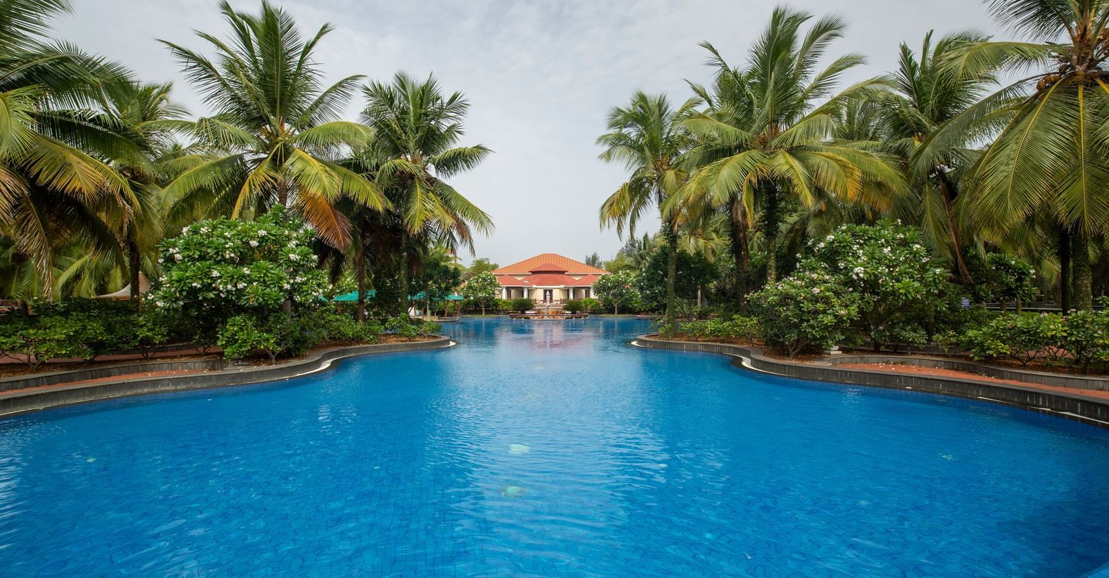 A beautiful swimming pool at a tropical resort, surrounded by palm trees and lush greenery and a building with a red tile roof in the distance - Ocean Spray, Pondicherry