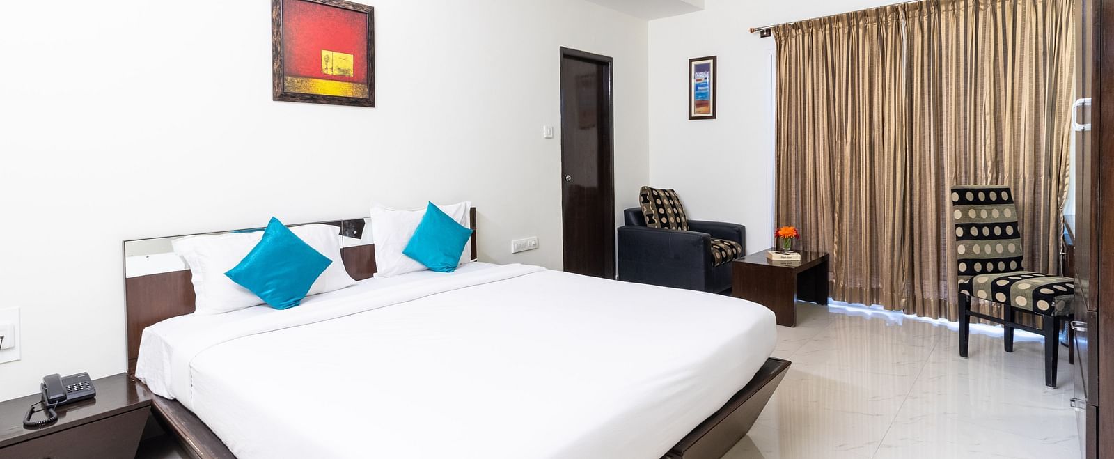 A room featuring a double bed with blue cushions and dark wooden furniture at Sanctum Suites Domlur, Bangalore.