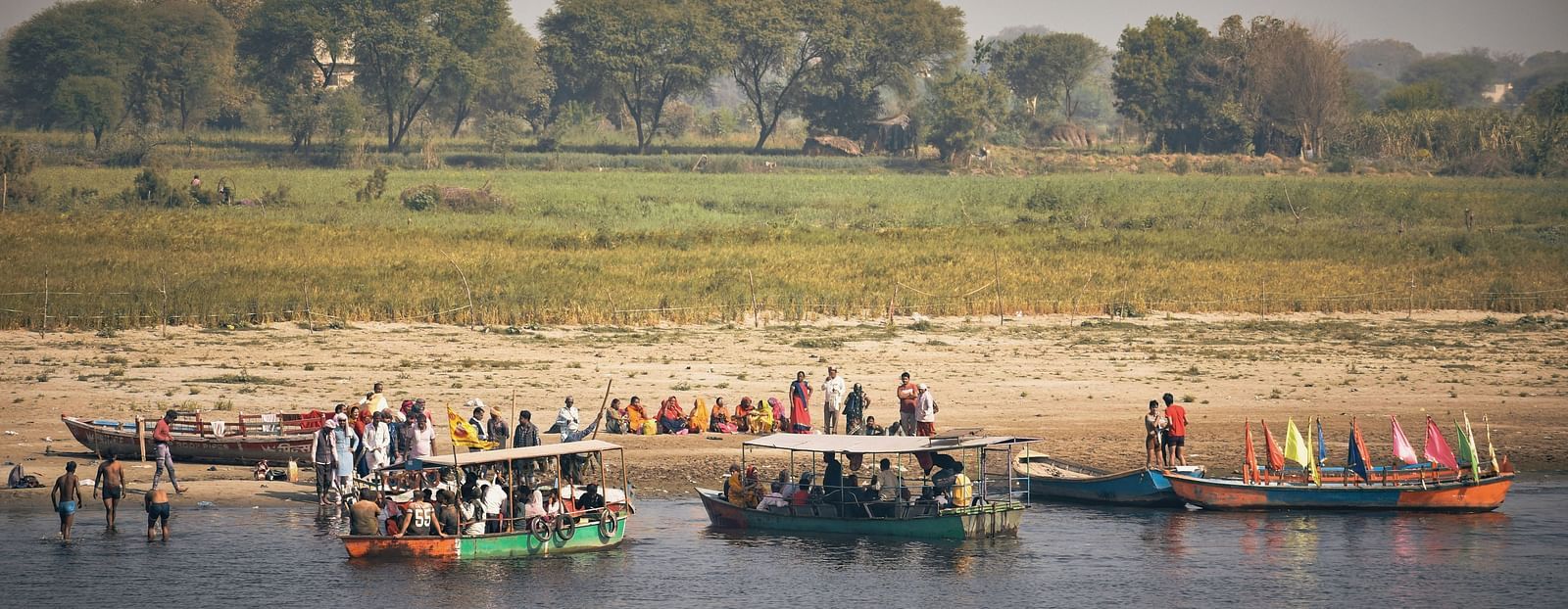 Empty boat and boats loaded with people on the bank of the river next to paddy field while other people are waiting on the bank for the boats to be vacated