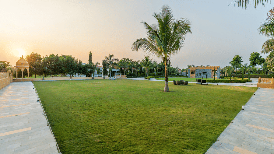 A view of the outdoor lawn at EsthereaRaj Leela, Ranakpur, at sunset, featuring palm trees and perimeter walkways.