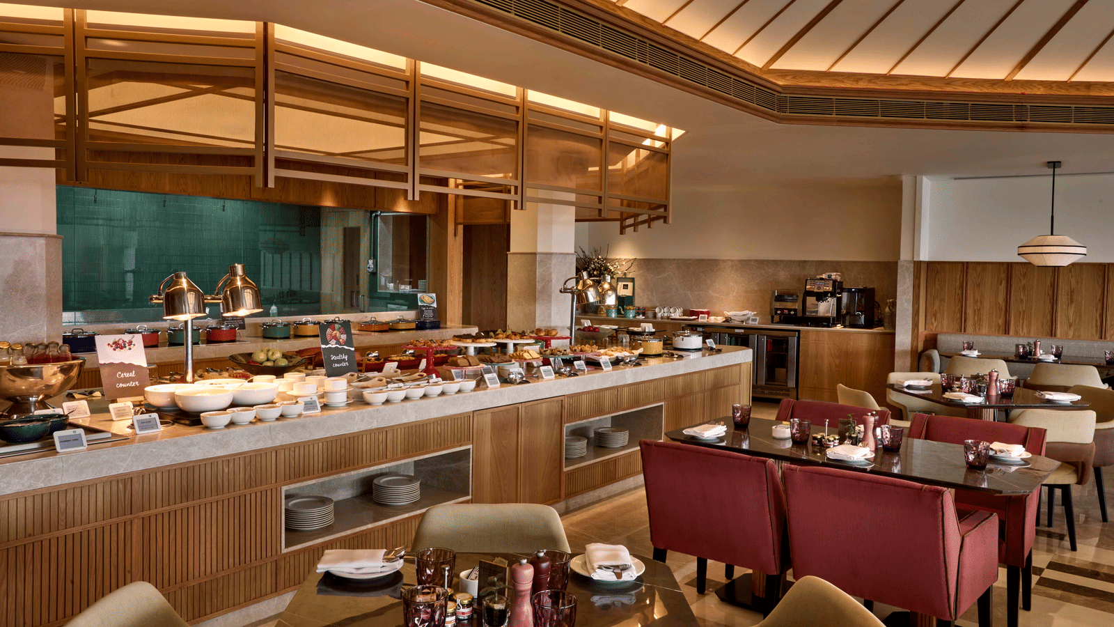 Warmly lit, modern restaurant dining room with a long buffet counter, wood panelling, and seating featuring maroon banquettes and beige chairs at Athiva Resort & Spa, Khandala - Best Restaurants in Khandala