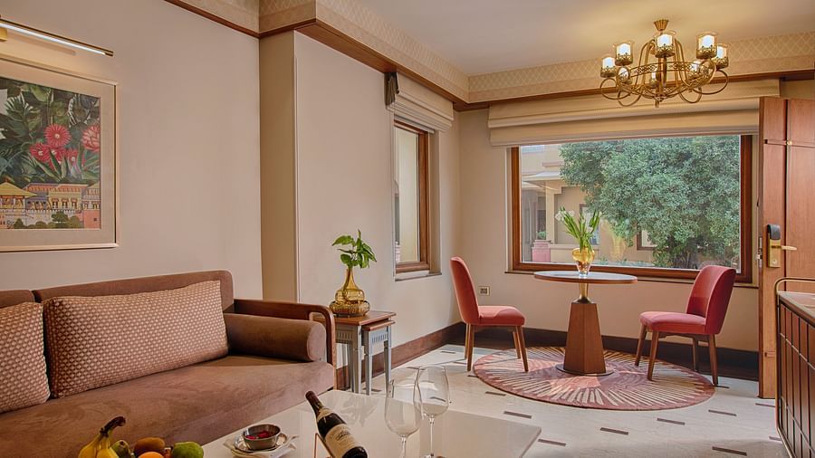 A bright and spacious living area with a comfortable sofa, a round dining table for two, elegant curtains, and a large window overlooking a garden