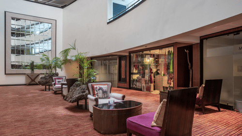 Hotel lobby of The Boma Nairobi with plush seating on patterned carpet. Large plants add ambiance. Glass storefronts line the wall, and a large mirror reflects geometric lines.