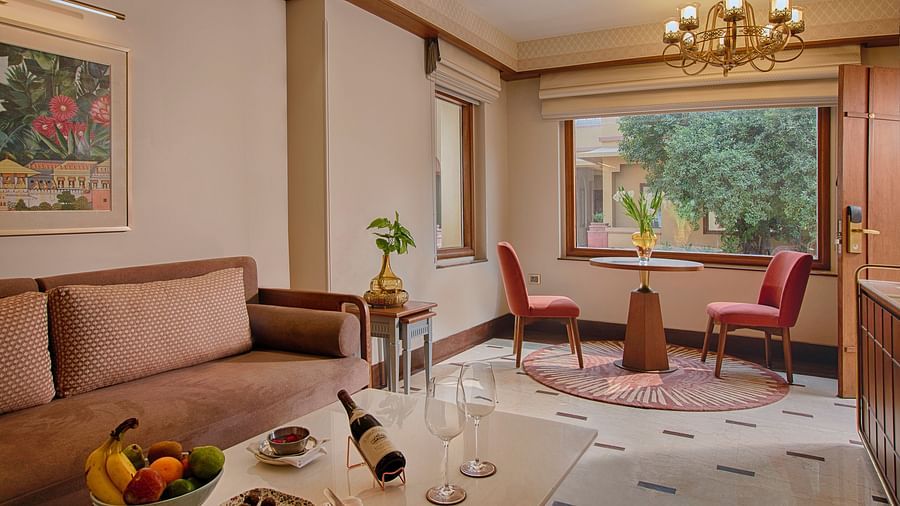 A bright and spacious living area with a comfortable sofa, a round dining table for two, elegant curtains, and a large window overlooking a garden