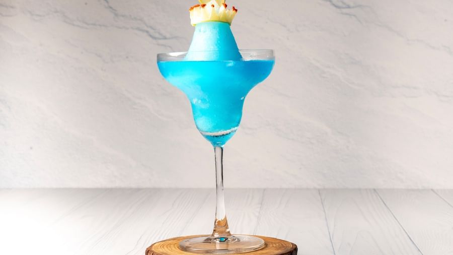 A frozen cocktail in a martini glass is garnished with a lemon slice and a red spice rim on a wooden coaster.