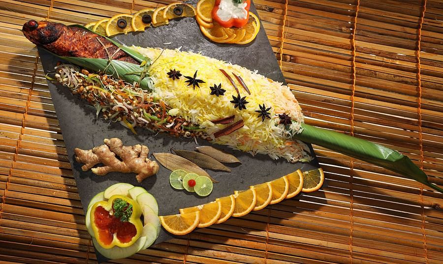 A decorated platter on a bamboo mat features a fish stuffed with yellow rice and garnished with star anise, sprouts, ginger, lime slices, and intricately cut orange and bell pepper pieces at UDS Group of Hotels.