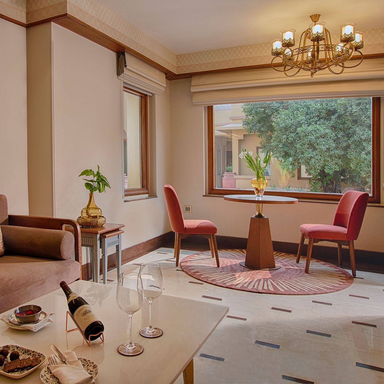 A bright and spacious living area with a comfortable sofa, a round dining table for two, elegant curtains, and a large window overlooking a garden