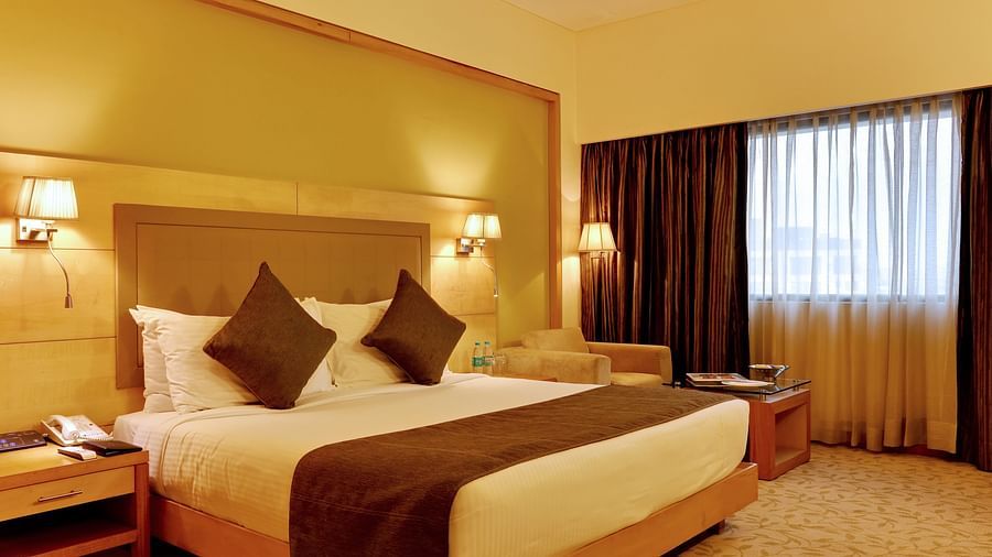 An image of a Deluxe Room with double size bed, bedside table and lam on either side and a huge window covered with curtains - The Suryaa New Delhi