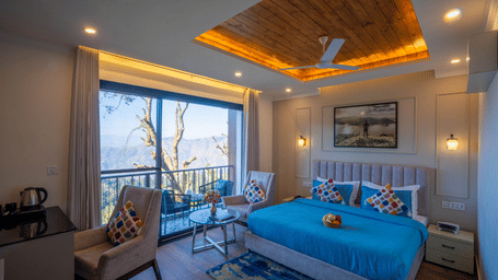 A bedroom with a king size bed, 2 chairs, large window, wooden ceiling panels, and exterior hill view at Perfectstayz Premium Westend Mall Road, Mussoorie.