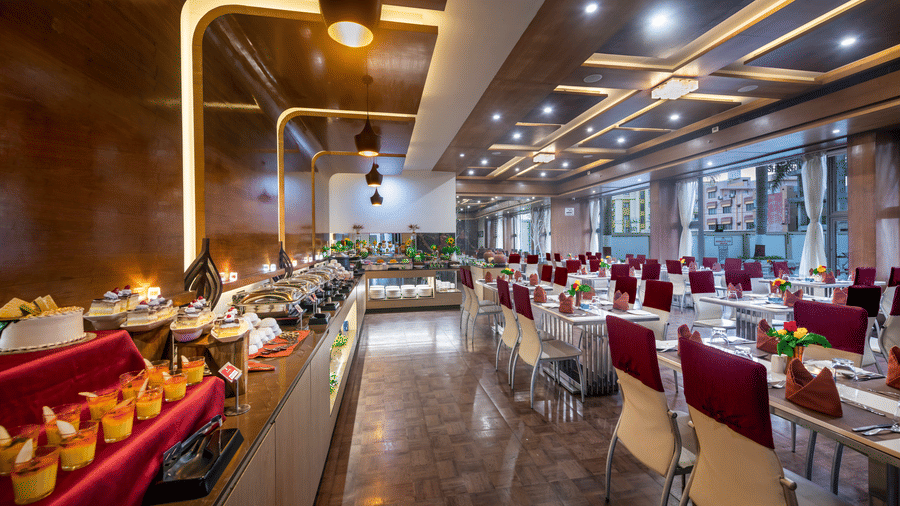 A restaurant with modern decor, food counters, and neatly set dining tables featuring a long counter displaying assorted desserts and dishes at Lords Inn Somnath