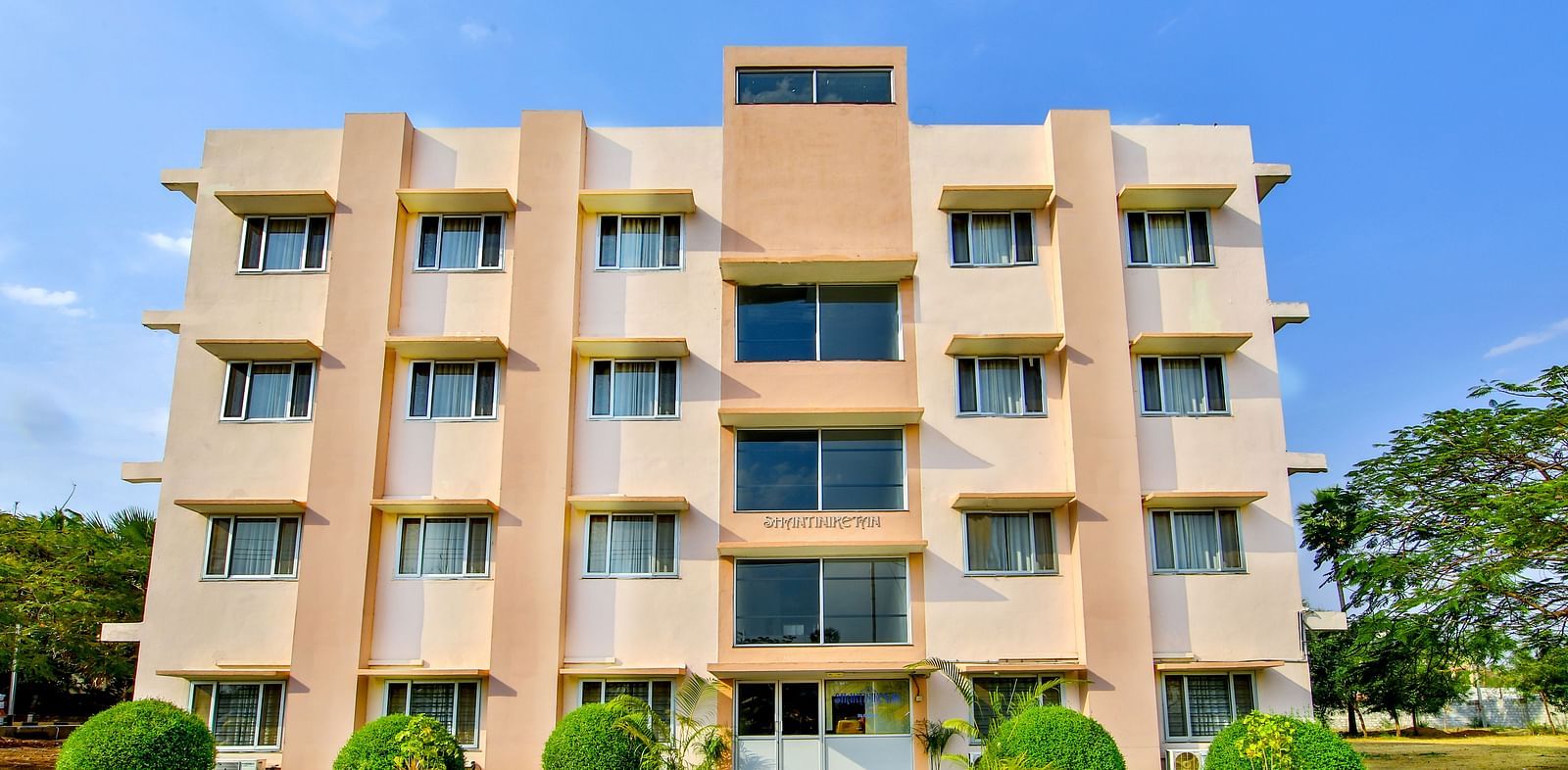 Four-story building with a pink and beige exterior, front view with a clear blue sky background at Greens Inn - Modern Hotel in Hyderabad.