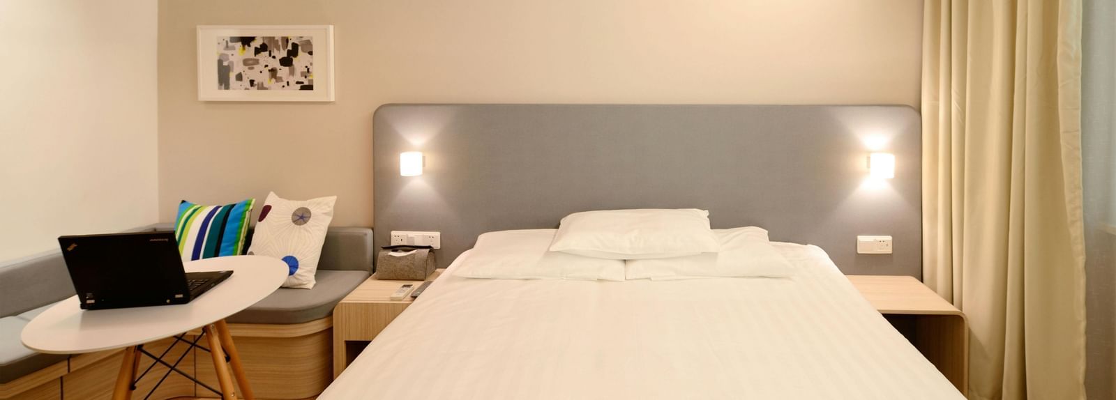 A bright and minimalist hotel room contains a bed with white linens, a desk with a laptop, and light-coloured curtains.