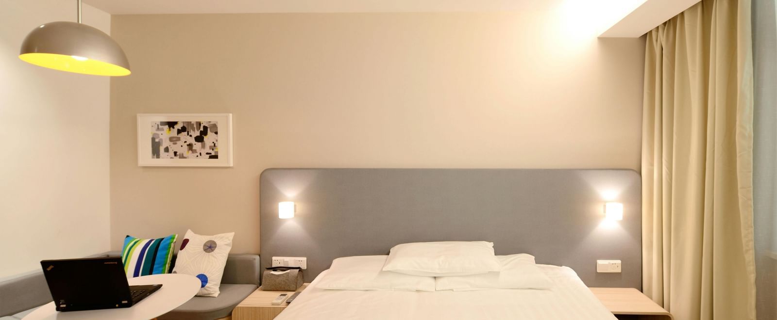 A bright and minimalist hotel room contains a bed with white linens, a desk with a laptop, and light-coloured curtains.