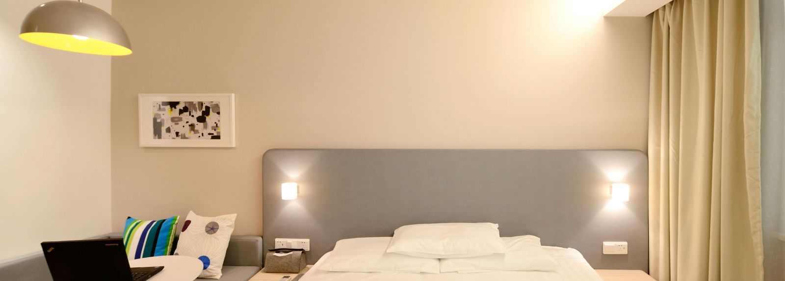 A bright and minimalist hotel room contains a bed with white linens, a desk with a laptop, and light-coloured curtains.
