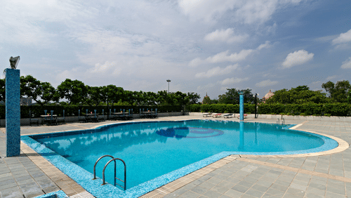 A luxurious outdoor swimming pool with crystal clear blue water surrounded by concrete decking, set against a backdrop of lush greenery and cloudy skies at a five star hotel in Lucknow - Clarks Avadh, Lucknow.