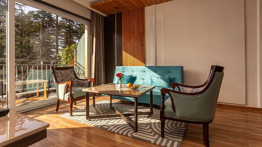 An image of a room with sofa set, centre table, and an expansive glass door through which rays of sun is gleaming inside - The Belvedere Himalayan Retreat, McLeodganj