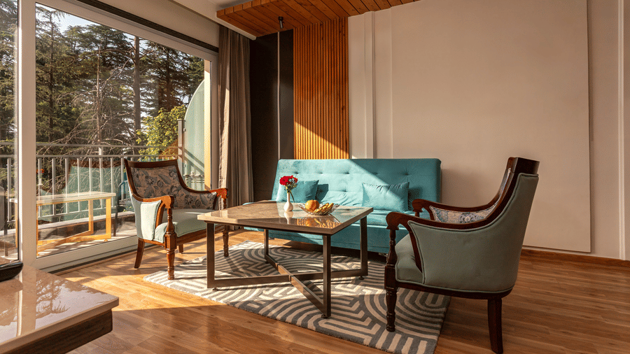 An image of a room with sofa set, centre table, and an expansive glass door through which rays of sun is gleaming inside - The Belvedere Himalayan Retreat, McLeodganj