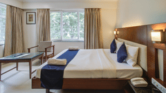Deluxe King room at Magnus Riverside Kalyani Nagar featuring a double size bed, a centre table and a chair. The room also have a telephone on bedside table and there are two big windows with curtains.