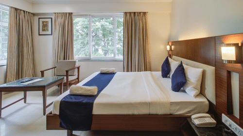 Deluxe King room at Magnus Riverside Kalyani Nagar featuring a double size bed, a centre table and a chair. The room also have a telephone on bedside table and there are two big windows with curtains.