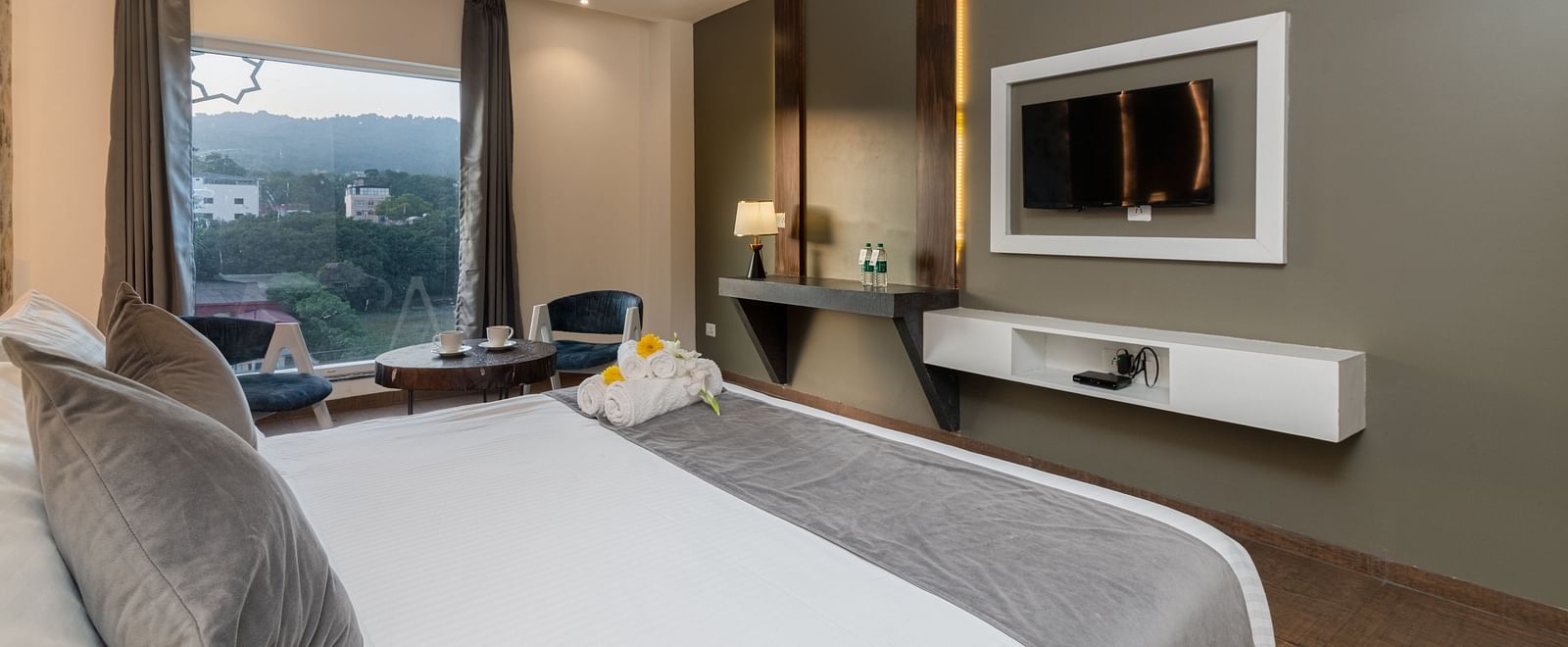 A room with a bed, a flat screen TV, two chairs and a coffee table, and an expansive glass window overlooking the view - DLS Ganga Bliss, Haridwar