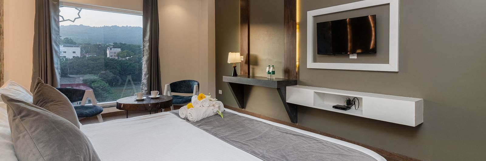 A room with a bed, a flat screen TV, two chairs and a coffee table, and an expansive glass window overlooking the view - DLS Ganga Bliss, Haridwar