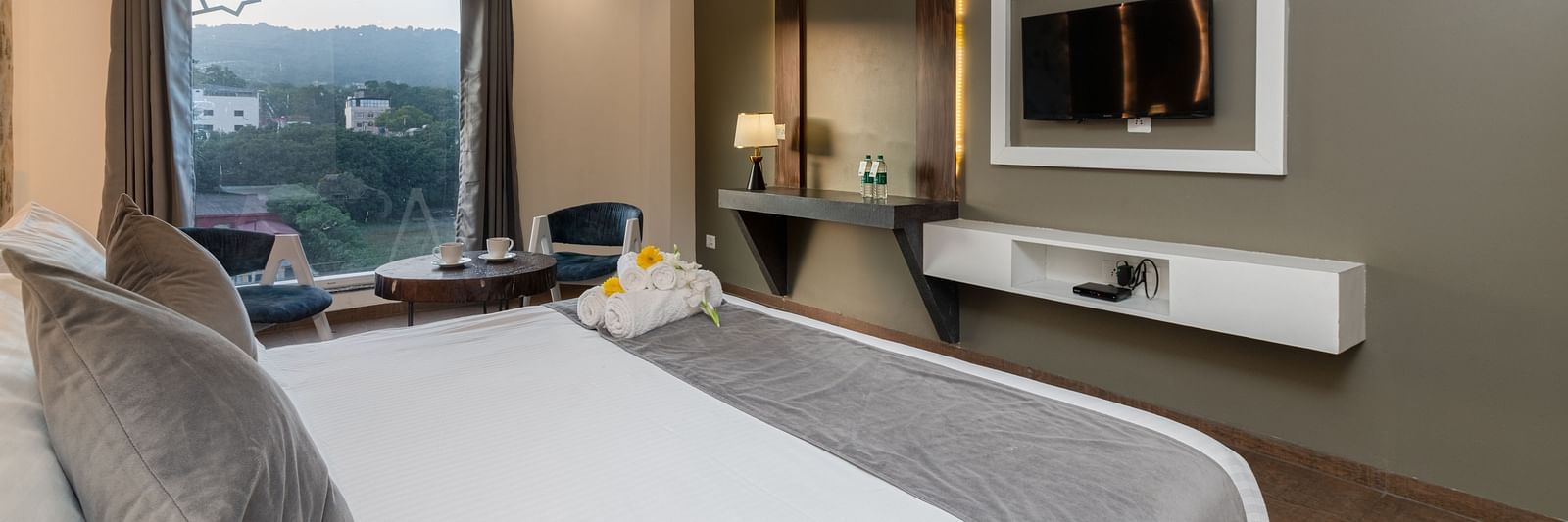 A room with a bed, a flat screen TV, two chairs and a coffee table, and an expansive glass window overlooking the view - DLS Ganga Bliss, Haridwar