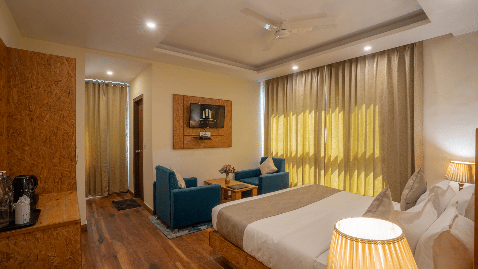 A hotel room, featuring a large bed with white linens, a small blue sofa, gold curtains, and a tall white bedside lamp | Perfectstayz Value Valley Vista