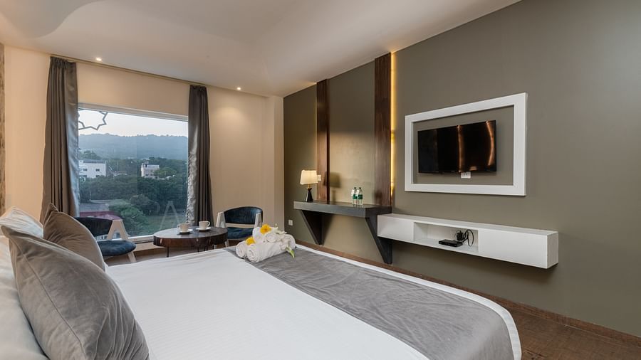 A room with a bed, a flat screen TV, two chairs and a coffee table, and an expansive glass window overlooking the view - DLS Ganga Bliss, Haridwar