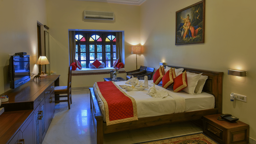 A bedroom at Garh Govind featuring a king size bed with a red runner, a side table, a television console, work desk, chair, and a window with grilles.