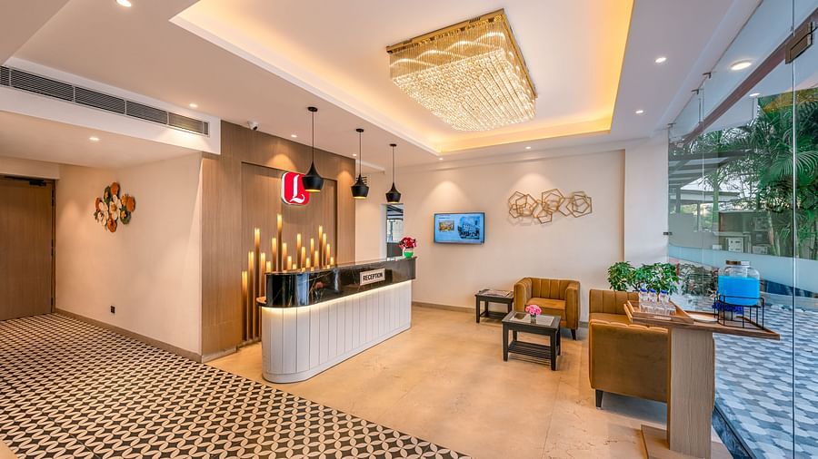 A hotel reception area with a front desk, chandelier, and comfortable seating. It boasts a patterned carpet, stylish modern furniture, and a scenic view through large windows - Lords Eco Inn Dehradun.