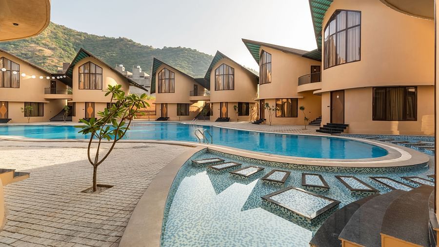 Exterior of The Qualia Resort Club & Brewery, Udaipur showcasing a swimming pool surrounded by buildings, reflecting the natural landscape.