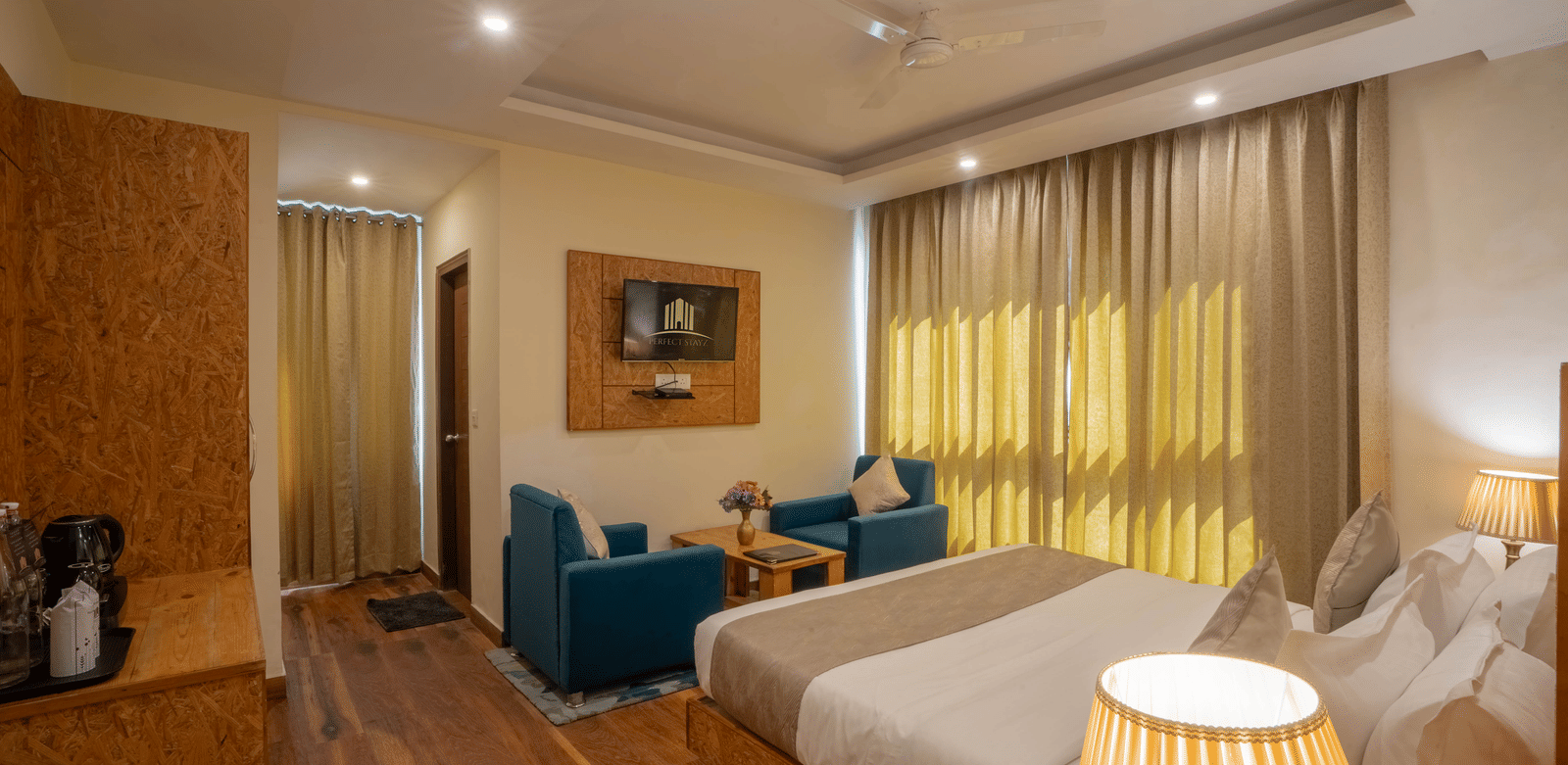 A hotel room, featuring a large bed with white linens, a small blue sofa, gold curtains, and a tall white bedside lamp | Perfectstayz Value Valley Vista
