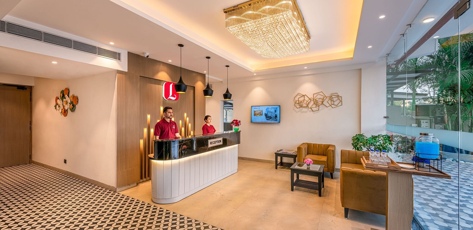 A hotel reception area with a front desk, two staff members, and a decorative chandelier. It features a patterned carpet, modern furniture, and a view through large windows - Lords Eco Inn Dehradun.