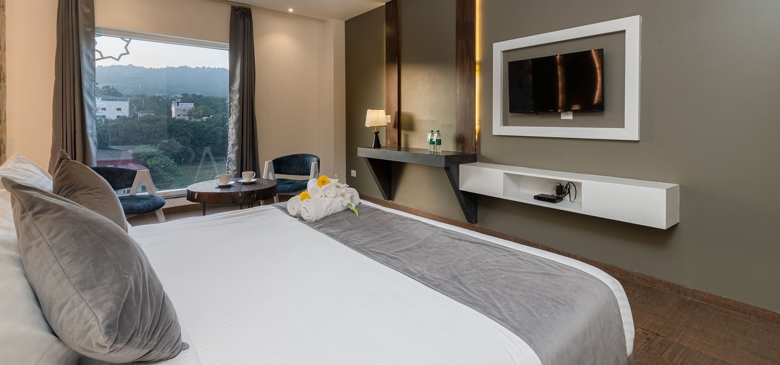 A room with a bed, a flat screen TV, two chairs and a coffee table, and an expansive glass window overlooking the view - DLS Ganga Bliss, Haridwar
