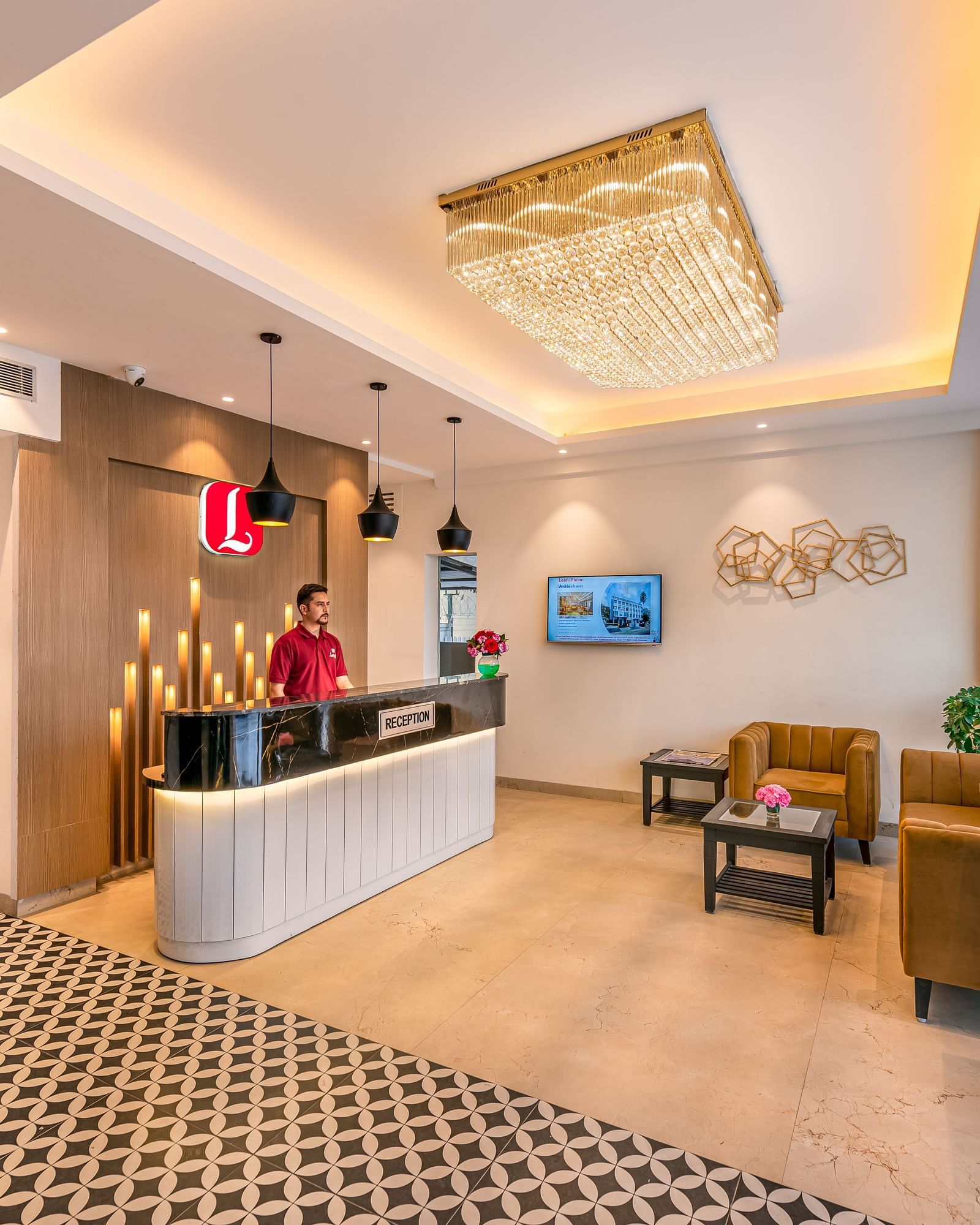 A hotel reception area featuring a front desk, a staff member, and a decorative chandelier. It includes a patterned carpet, modern furniture, and a view through large windows - Lords Eco Inn Dehradun.
