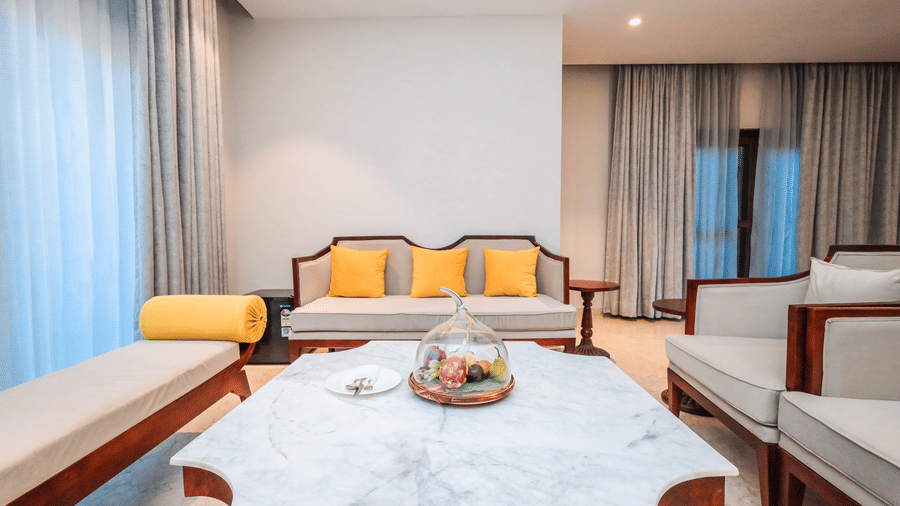 A room shows a centre table with fruit, long seating on both sides, cushions on the seats and curtains on a window at Ananta Spa & Resort, Jaipur.