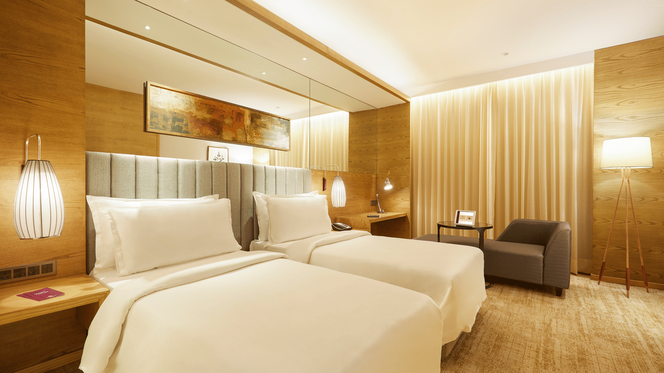 The Premier Room at Residency Towers, Chennai showcasing a twin beds, wooden flooring, side table with lamps, and an armchair near glass window.