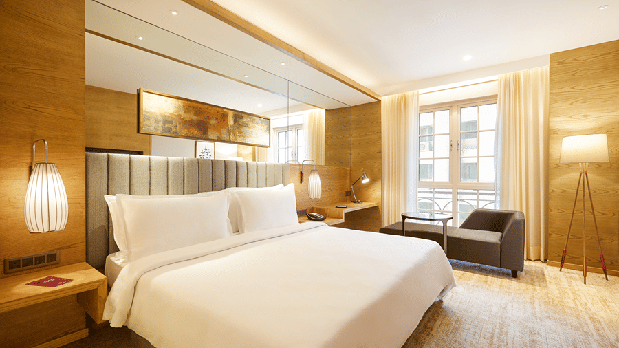 The Premier Room at Residency Towers, Chennai showcasing a double bed, wooden flooring, side table with lamps, and an armchair near glass window.