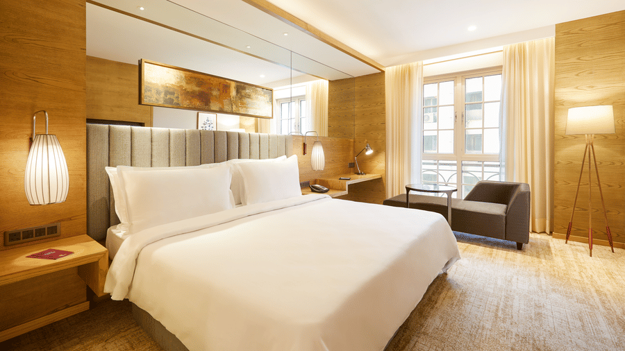 The Premier Room at Residency Towers, Chennai showcasing a double bed, wooden flooring, side table with lamps, and an armchair near glass window.