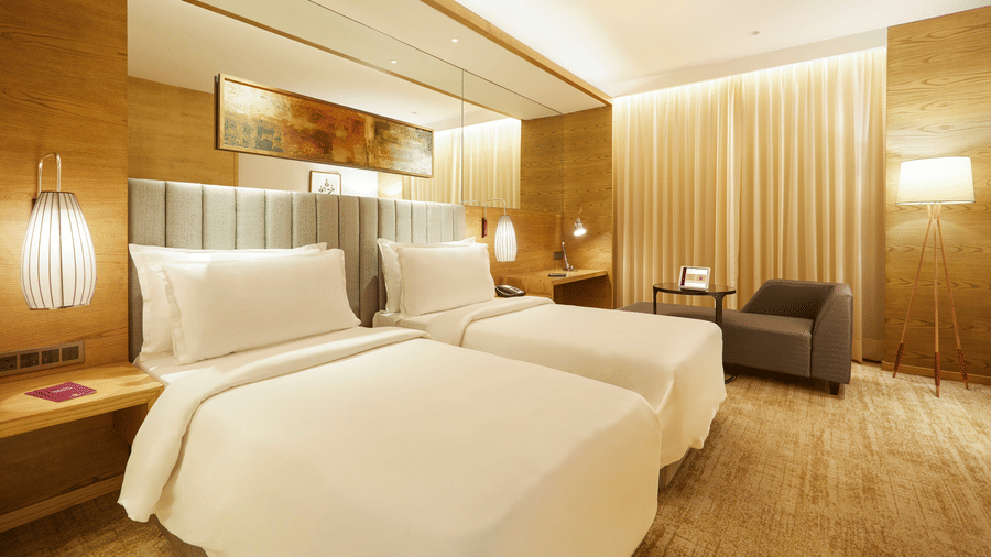 The Premier Room at Residency Towers, Chennai showcasing a twin beds, wooden flooring, side table with lamps, and an armchair near glass window.