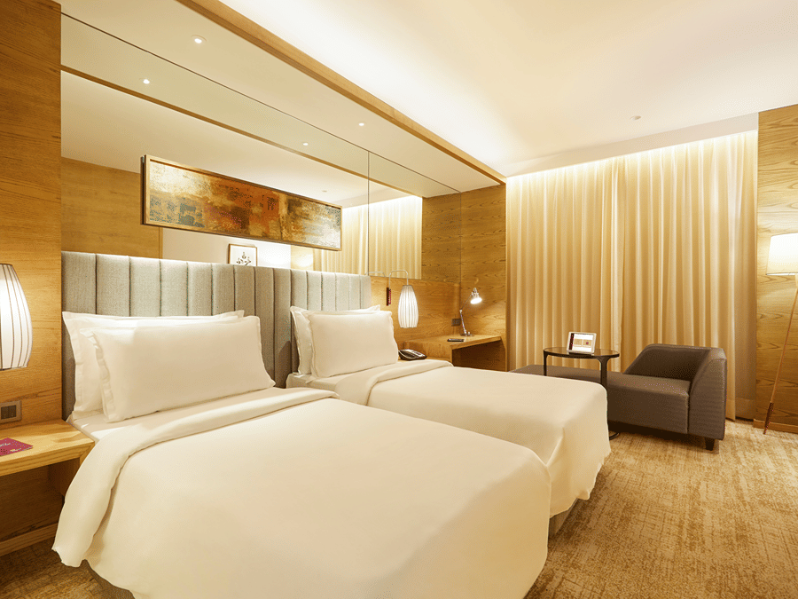 The Premier Room at Residency Towers, Chennai showcasing a twin beds, wooden flooring, side table with lamps, and an armchair near glass window.