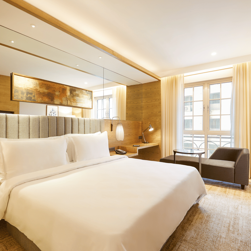 The Premier Room at Residency Towers, Chennai showcasing a double bed, wooden flooring, side table with lamps, and an armchair near glass window.