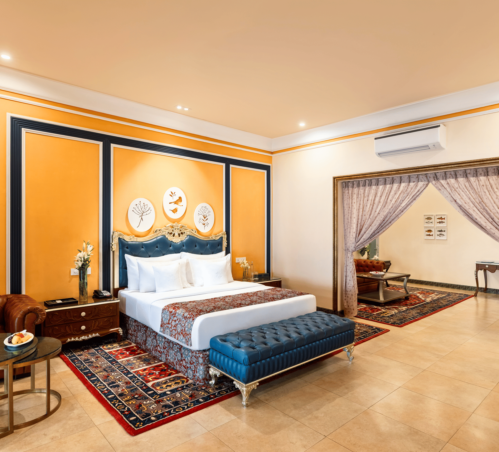 Suite with a king bed, a tufted bench, a leather sofa, coffee tables and an adjoining room through a gilded archway at MAYFAIR Palm Beach Resort, Gopalpur-on-Sea.