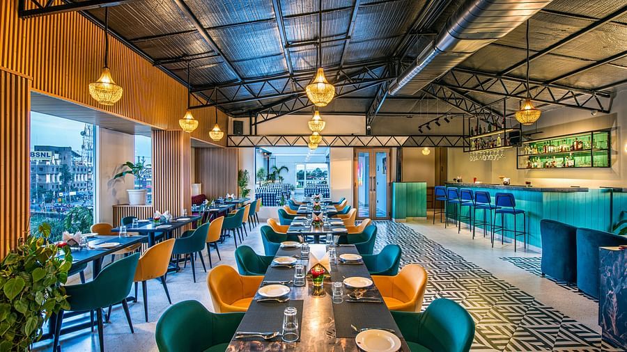 Restaurant interior with colourful chairs, dark tables, and hanging pendant lights. It features a high ceiling with exposed beams, a tiled floor, and a bar area in the background - Lords Eco Inn Dehradun.