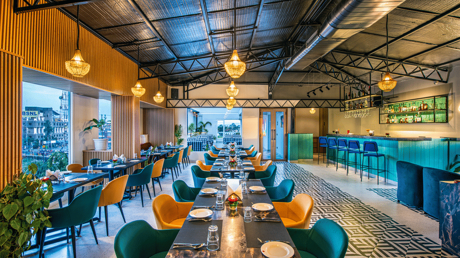 A view of the rooftop restaurant with colourful chairs and wooden tables, hanging pendant lights, high ceiling with exposed beams, a tiled floor, and a bar area in the background at Lords Eco Inn Dehradun.
