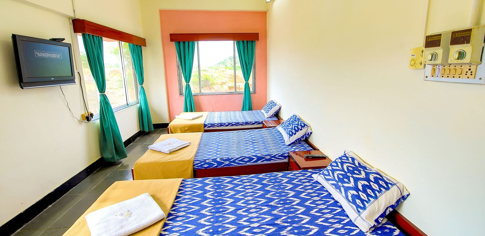 A side view of three beds with bedding in a room with a window and TV at Hotel Sahara - Comfortable Accommodations in Ramoji Film City, Hyderabad.
