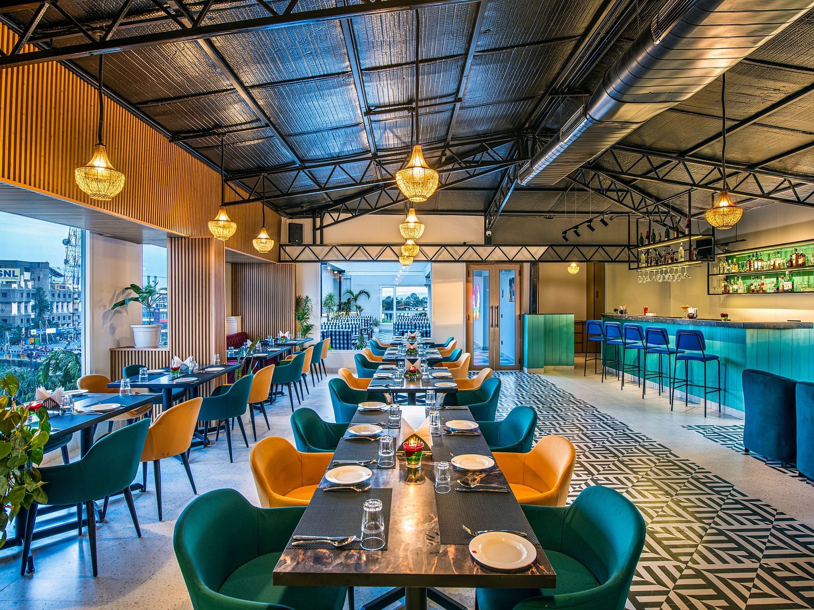 Restaurant interior with colourful chairs, dark tables, and hanging pendant lights. It features a high ceiling with exposed beams, a tiled floor, and a bar area in the background - Lords Eco Inn Dehradun.
