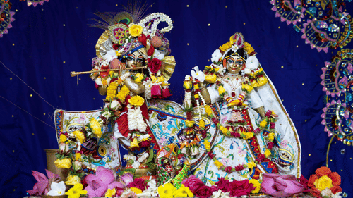 Intricately dressed statues of Hindu deities Krishna and Radha adorned with flowers and jewellery against a cloth backdrop.