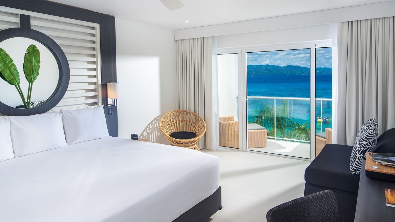 An image of a hotel room with a large bed, a cushioned chair and a balcony offering views of the sea - S Hotel Montego Bay, Jamaica