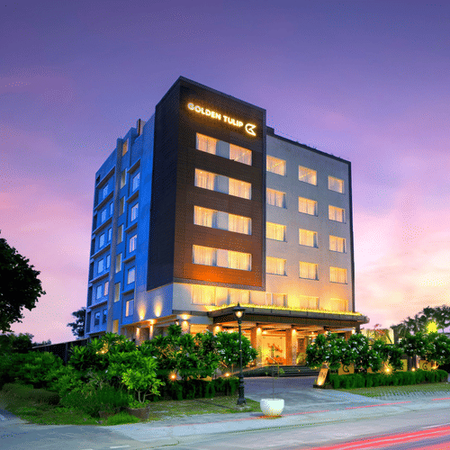 An image of the hotel building illuminated with bright yellow lights and a pink coloured sunset in the background - Golden Tulip, Agra